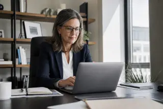 Confident woman in her fifties working on a laptop in a bright, modern office, focused and professional in a documentary photography style.