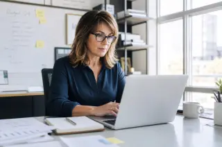 Confident woman in her 50s working focused at a laptop in a modern, well-lit office environment, reflecting active and professional engagement.