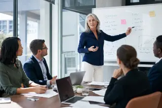 A confident 60-year-old woman leads a team meeting in a modern, bright office with attentive colleagues of varying ages.