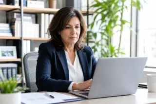 Woman in her 50s focused on a laptop in a modern, sunlit office, appearing professional and engaged in her work.