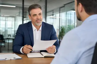 A confident 57-year-old man in a bright, modern office engaged in a professional conversation with a younger colleague, both focused and attentive amid contemporary workspace décor.