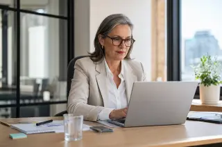 Senior woman, aged around 55, working intently on a laptop in a sunlit, modern office, exuding professionalism and confidence.